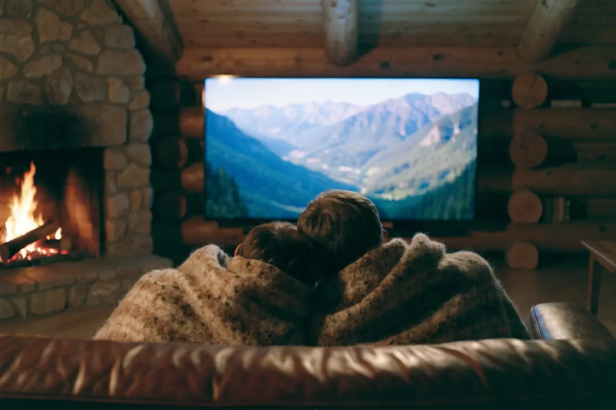A couple under a blanket watching a big-screen TV beside a cabin fireplace