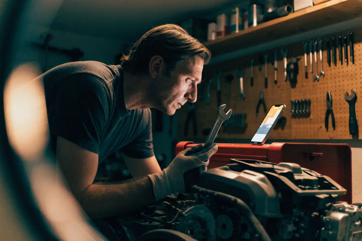 A man in his garage, wrench in hand, following a how-to video on his phone